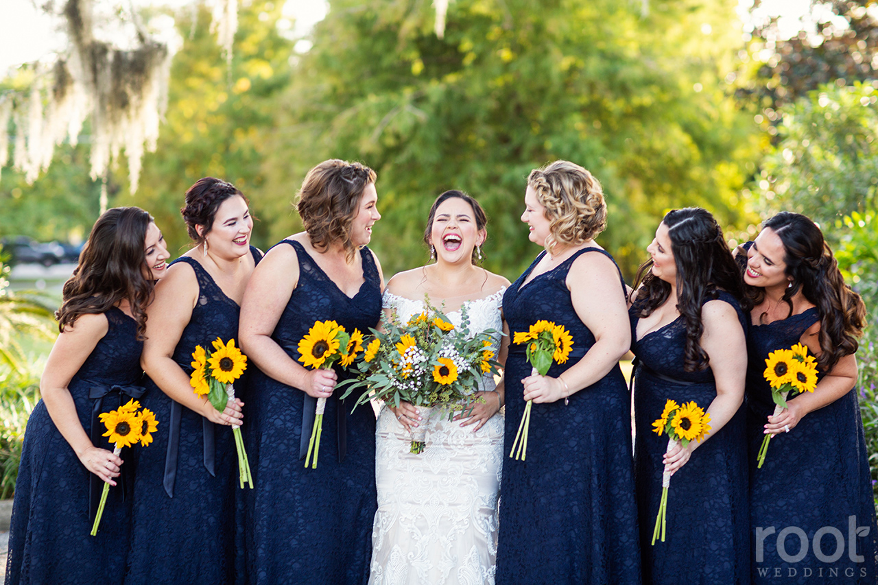 A bride laughing with bridesmaids in navy dresses with sunflower bouquets